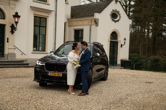 Bruidsstel poseert bij een zwarte auto voor een elegante villa.