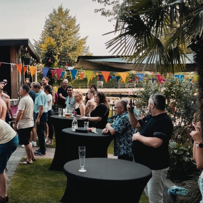 Buitenevenement met mensen, tafels en feestdecoraties in een groene omgeving.