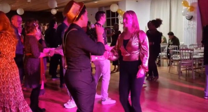 Twee mensen dansen en praten op een feest, omringd door andere feestgangers.
